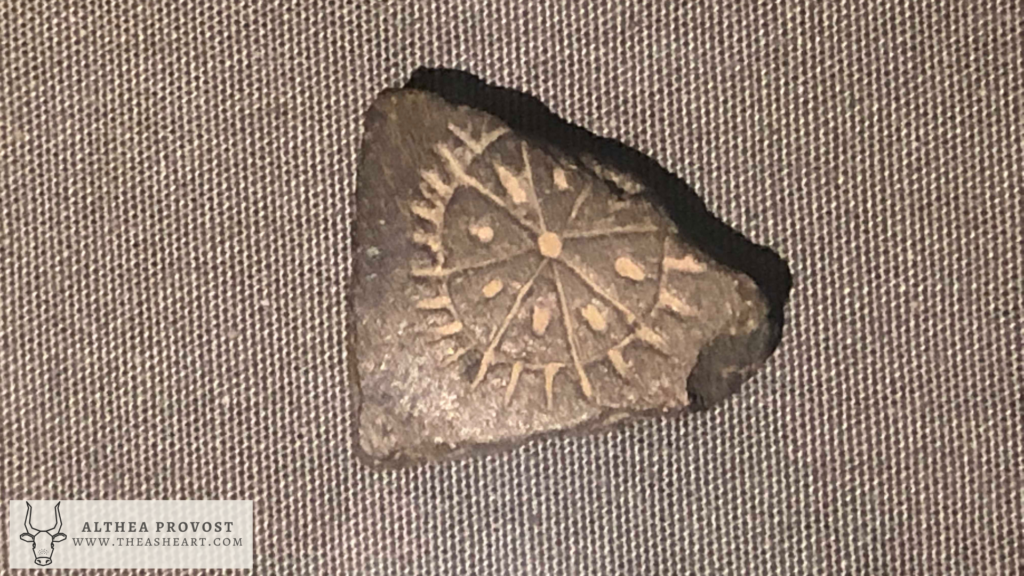 Triangular stone fragment engraved with a circular sun‑like motif featuring a central dot and radiating lines.