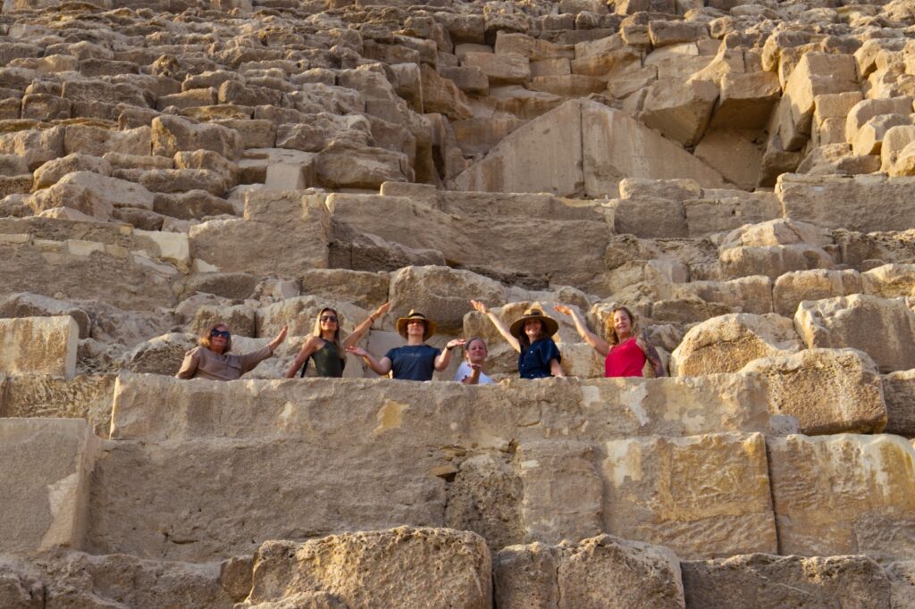 Chevron‑Shaped Structure during Starseed Egypt Adventure 2024 with Althea Provost, partially obscured behind the group standing on the north‑face blocks of the Great Pyramid, with six participants raising one arm in alignment with the pyramid slope