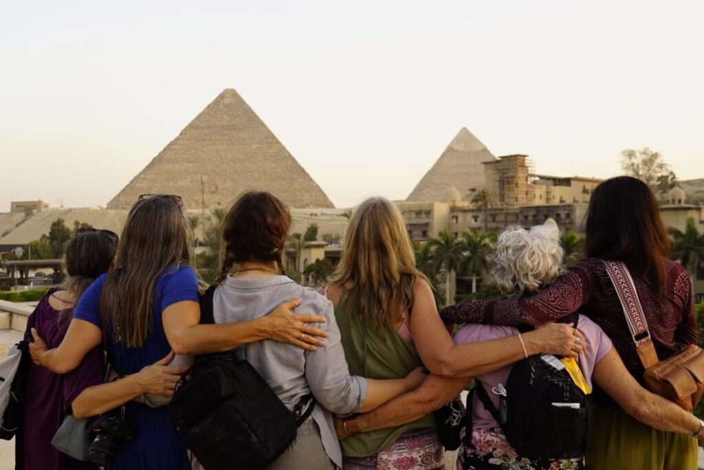 Chevron‑Shaped Structure during Starseed Egypt Adventure 2022 with Althea Provost, viewed from the rooftop of the four‑story Marriott Mena House Hotel, with seven participants standing arm‑in‑arm facing the Great Pyramid and Pyramid of Khafre