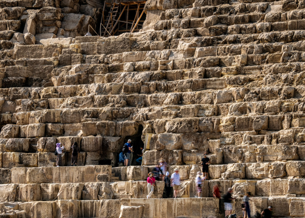 Crowds of tourists climbing partway up the Great Pyramid’s north face, with a visible entrance cavity and scaffolding set among the massive limestone blocks