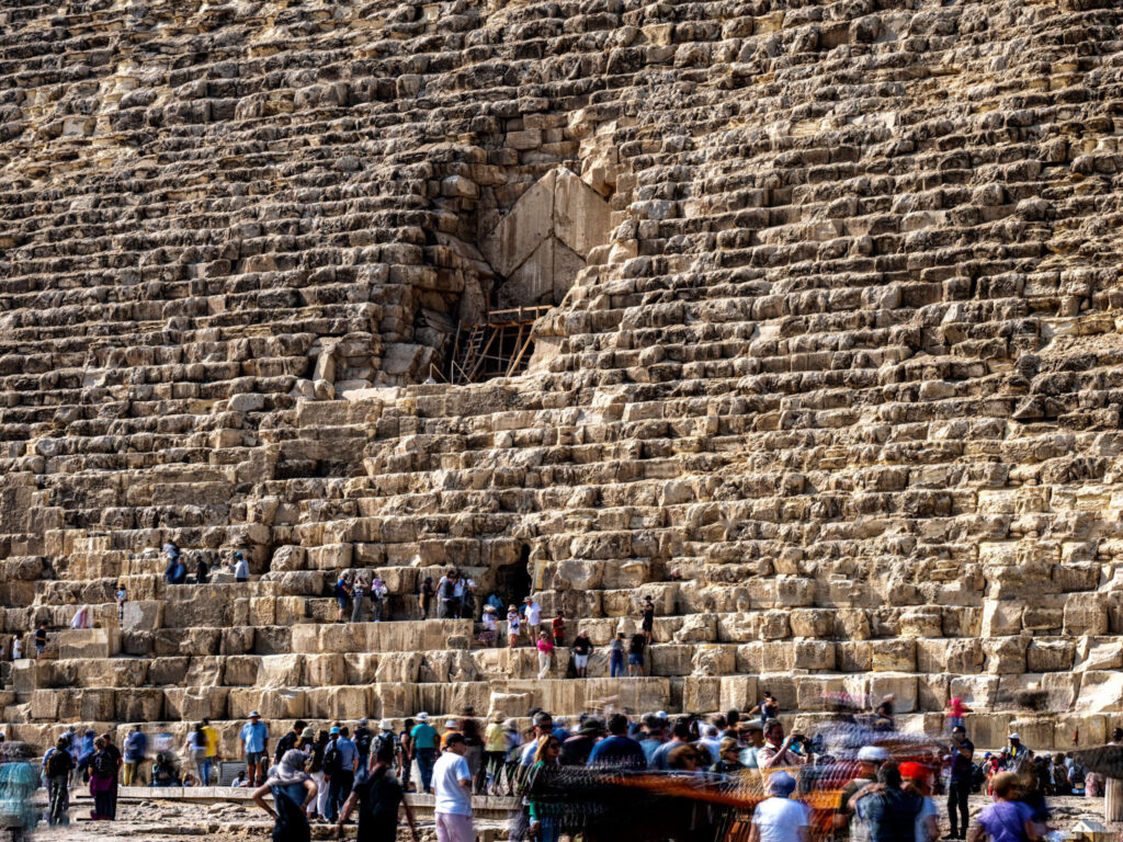 Chevron‑shaped structure above the Great Pyramid’s north entrance with tourists standing and climbing on the limestone blocks and scaffolding surrounding the access opening