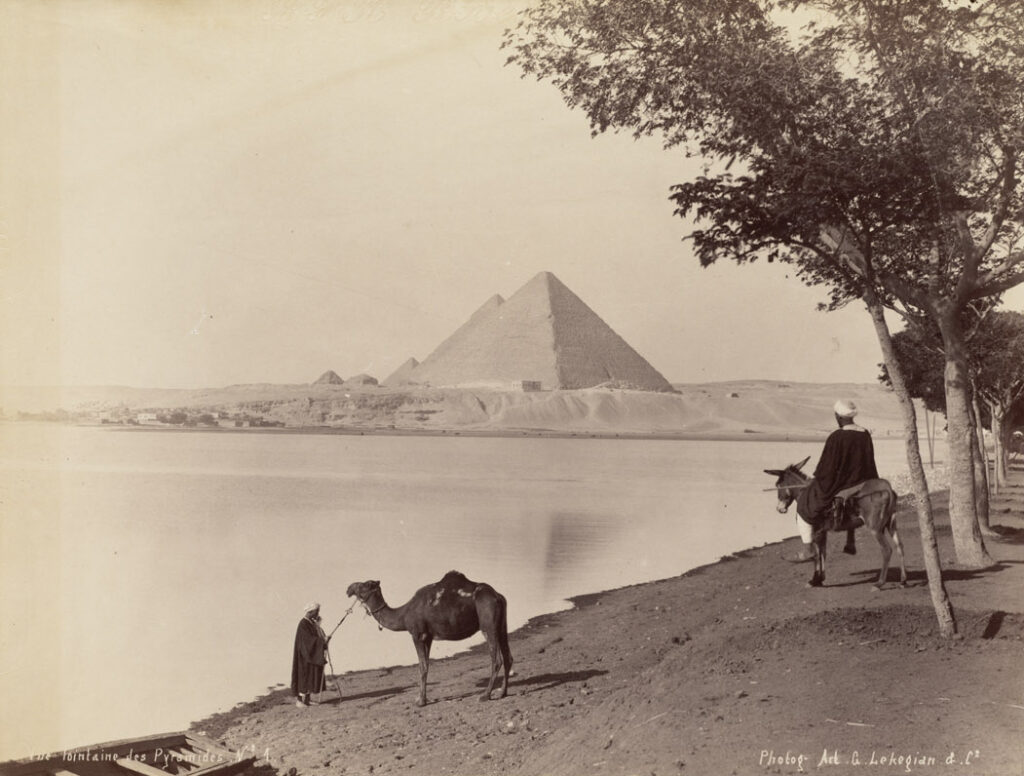 Distant view of the pyramids with a man beside a camel and another seated on a donkey under trees in Lekegian photograph No. 1