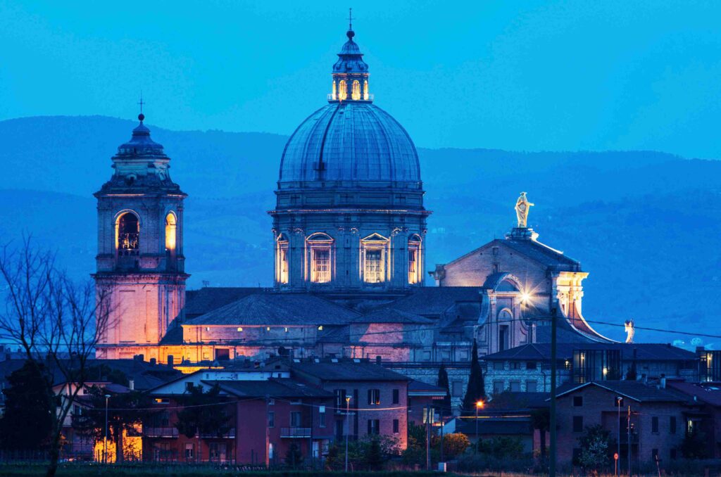 Saint Francis Walking Tour with Althea Provost. Dramatic dusk view of a domed cathedral with illuminated windows, flanked by a smaller bell tower and residential buildings in the foreground.