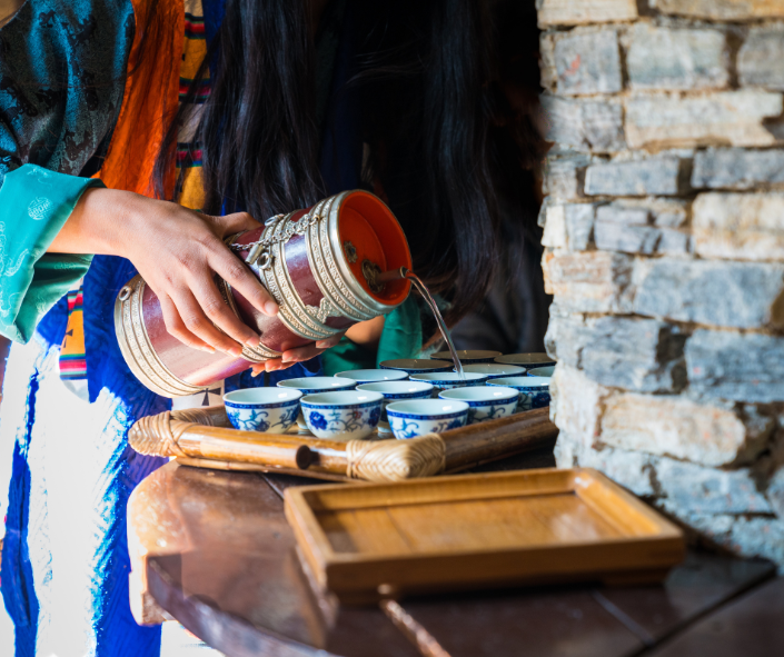 Starseed Bhutan Adventure with Althea Provost — a Bhutanese woman in traditional dress pours warm butter tea into blue‑and‑white cups inside a rustic stone‑and‑wood setting, welcoming guests with cultural hospitality.